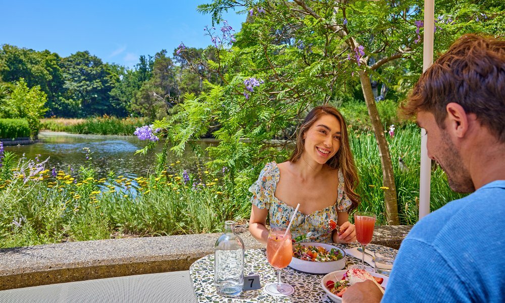 A couple dining on colourful brunch dishes and cocktails beside a lake in a green park.