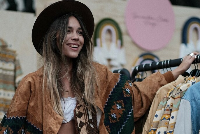 Smiling bohemian woman with long hair and black hat, standing next to a rack of Southwestern clothing.