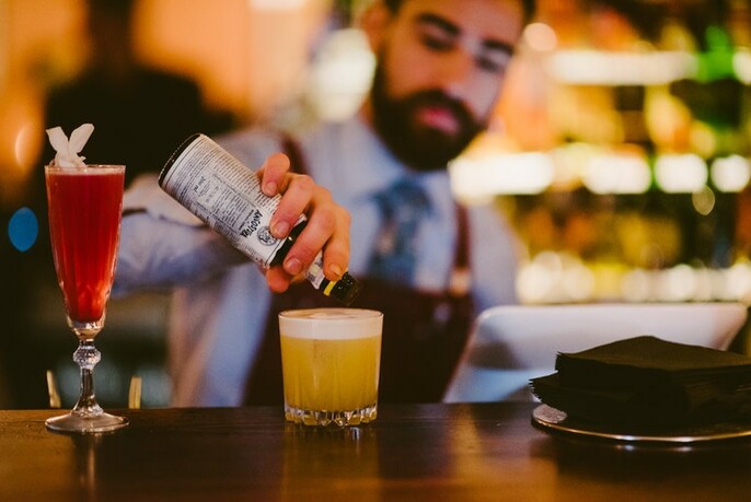 Bearded bartender pouring bitters into a yellow cocktail on a wooden bar, alongside a long red cocktail glass.