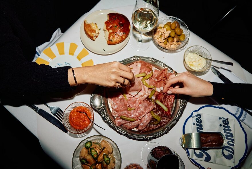 Two hands sampling cold meats from a charcuterie board, with surrounding appetisers and plates with cutlery.