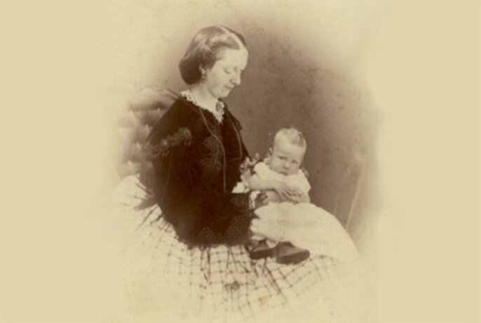 Vintage sepia-toned photograph of a woman holding a baby on her lap, in a vignette style.