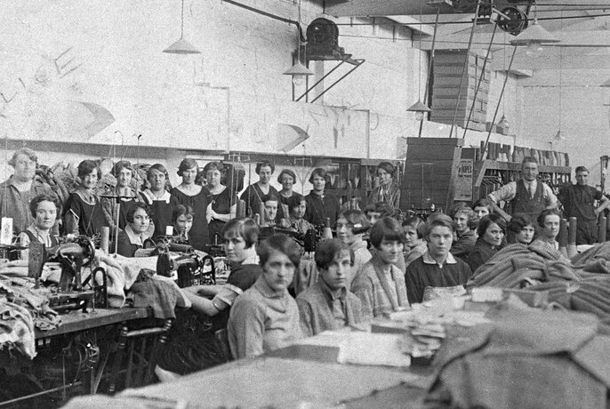 Vintage black and white image of many women sitting in a garment factory warehouse, looking seriously at the camera, seated behind long tables or at sewing machines.