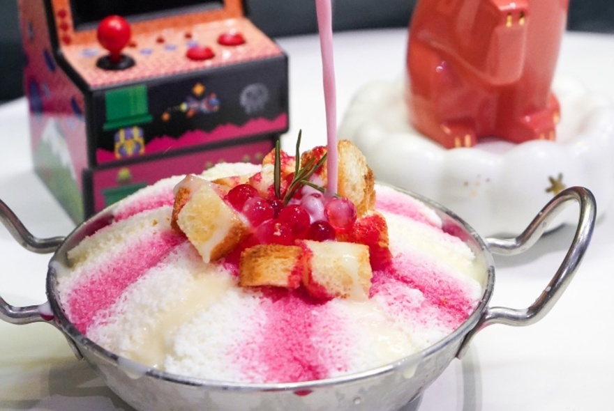 Shaved ice dessert in large stainless steel bowl with handles, with pink topping being poured on it.