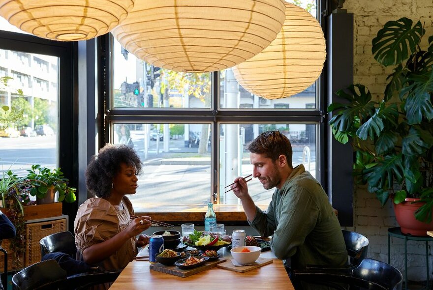 A couple eating a meal under large lantern lights in a sunny cafe.