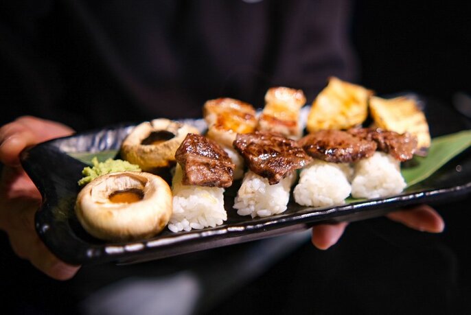 Hands holding a  dark plate of small food morsels, including small barbequed pieces of meat on rice.