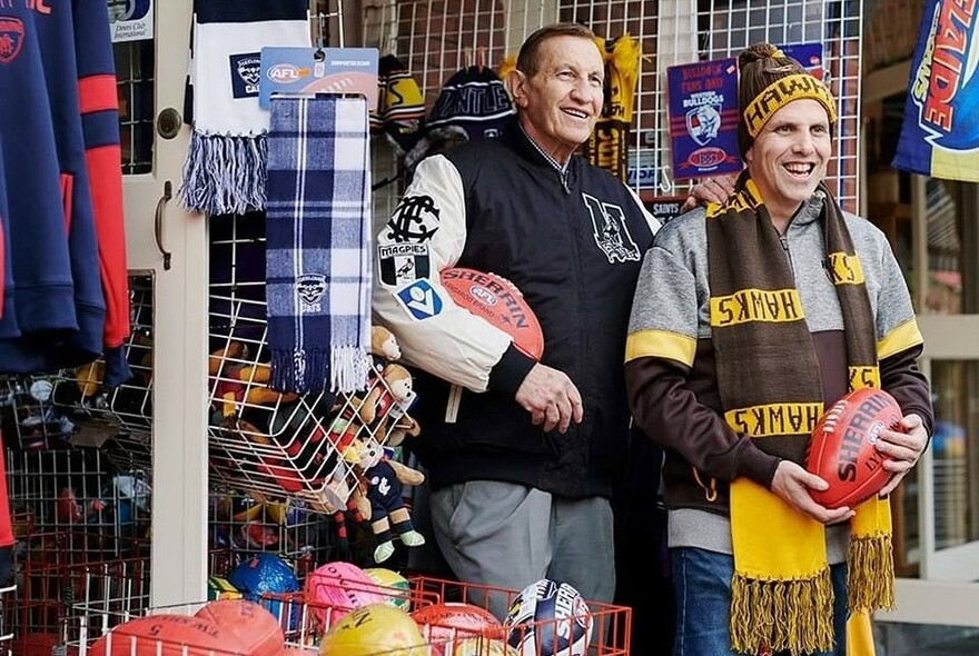Two people wearing sporting merchandise, holding footballs and standing in front of a stall selling sporting goods.