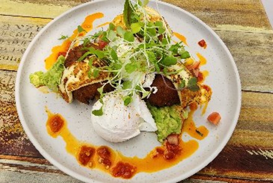 A breakfast plate featuring poached eggs and smashed avocado with a red sauce and microgreens.