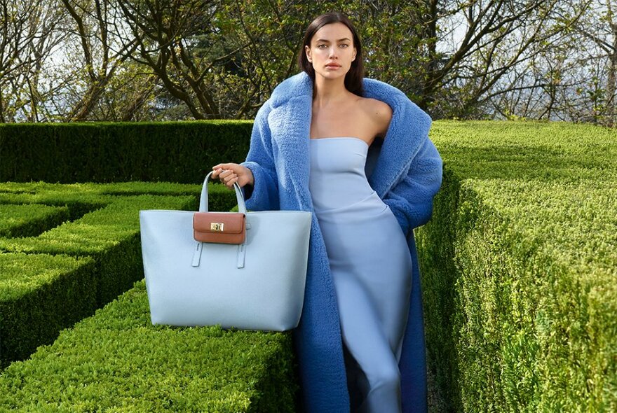 Model wearing a body hugging pale blue strapless dress and blue coat, carrying an oversize pale blue leather tote bag, standing in a well manicured low hedge maze.