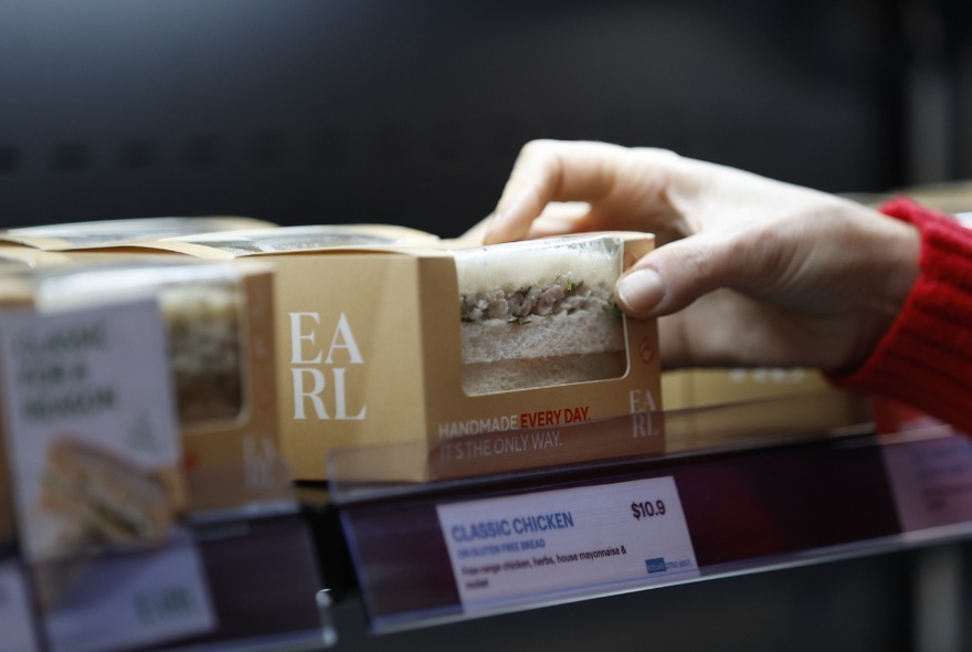 A hand selecting a sandwich in a cardboard box off a shelf with a pricing label below.