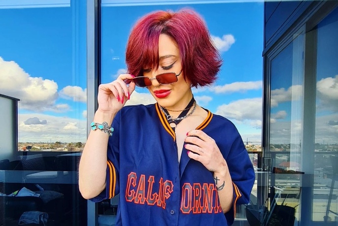 Woman with short, straight pink hair, lowering sunglasses and looking down, in short-sleeved blue bomber jacket, reflection of cityscape in windows behind.