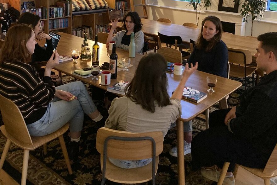 People seated around a table, with books and bottles of wine before them, in a room, conversing in soft lighting, a shelf of books against one wall, plants and other tables and chairs in the background.