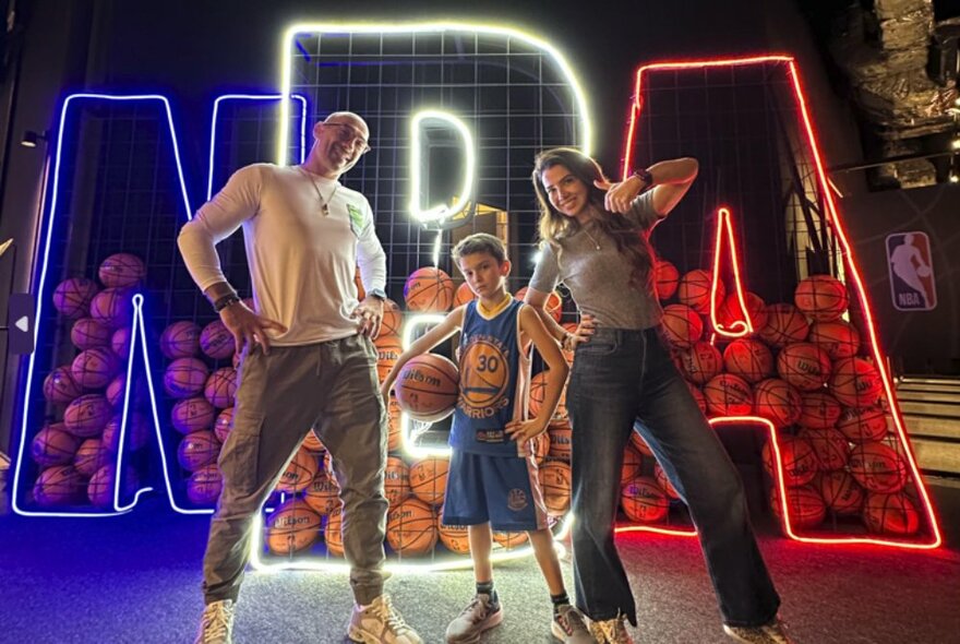 A tall man and woman posing next to a boy in NBA uniform and holding a basketball, in front of a neon NBA sign filled with basketballs.