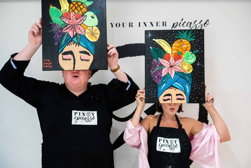 Two people holding up paintings of Carmen Miranda in front of their faces.