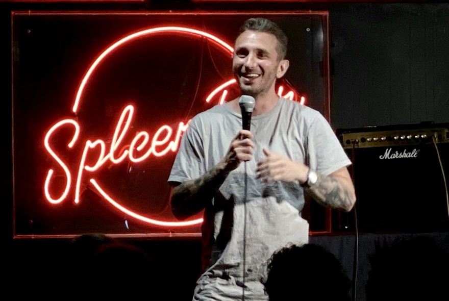 Stand-up comedian Tommy Little on stage in front of Spleen neon sign, holding microphone and smiling.