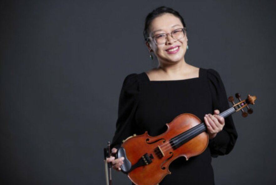 Violinist Tiffany Cheng, wearing a black dress and holding her violin, posed against a dark grey background.