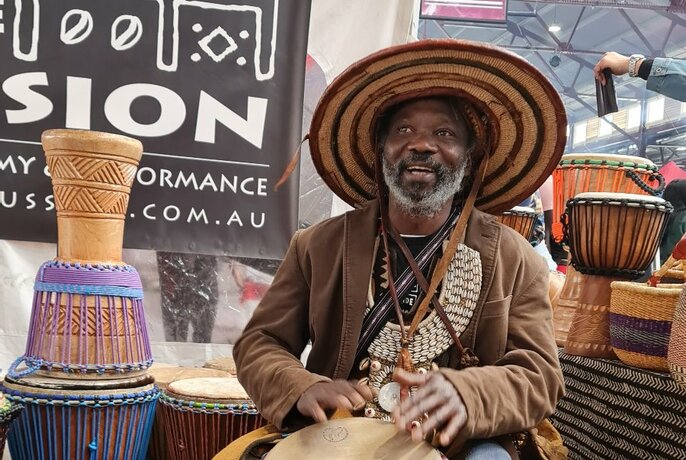 Seated smiling man wearing a hat and beads,  tapping his hands on a drum, with upturned samll african style drums surrounding him. 