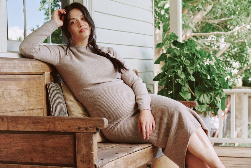 Pregnant woman in a stretchy neutral-coloured dress sitting on a wooden bench seat on a house verandah.