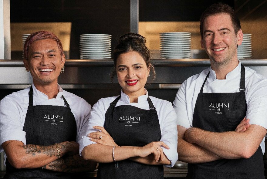 Chefs Kishwar Chowdhury, Callum Hann, and Khanh Ong, all from previous seasons of the TV show MasterChef, wearing matching chef's aprons, standing in a commercial kitchen.