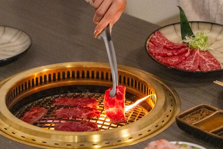 Hands holding tongs and cooking marbled beef on a gold-rimmed Japanese tabletop barbecue.