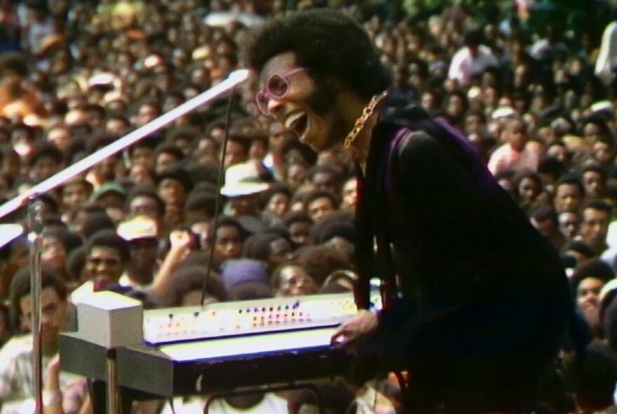 Sly Stone performing at a keyboard and microphone in front of a large seated audience in a still from the film Summer of Soul.
