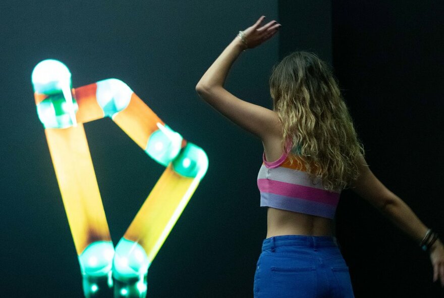 A person with their arms in the air, back to us, seemingly interacting with stick-shaped coloured lights on a wall.