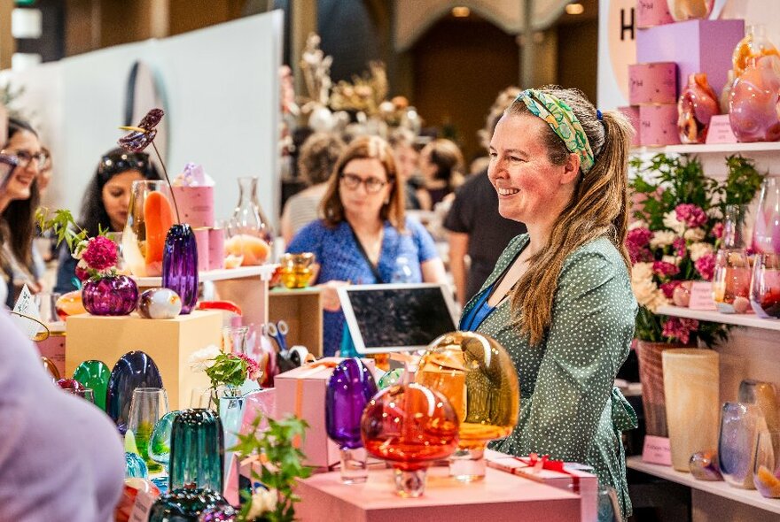 People browsing a designer stall displaying coloured glassware, and talking to the smiling stallholder inside the Royal Exhibition Buildings.