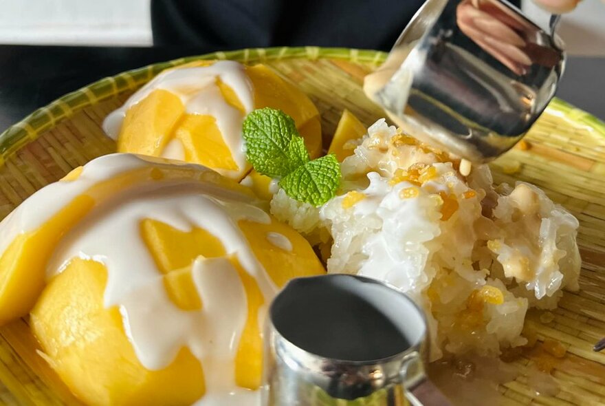 Silver sauce jug pouring white cream over dish of lemon Asian dessert.