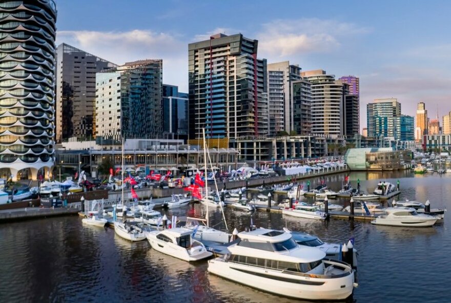 Docklands marina with boats moored by the walkway and city towers in the background.