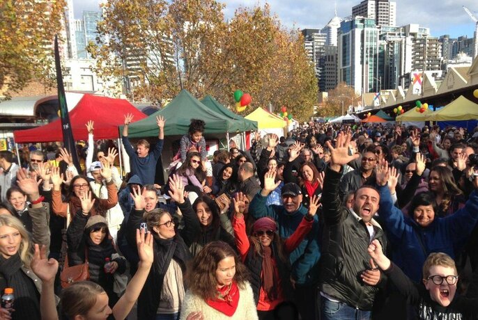 Large crowd of people outdoors at Queen Victoria Market enjoying themselves, tall city buildings in the background.