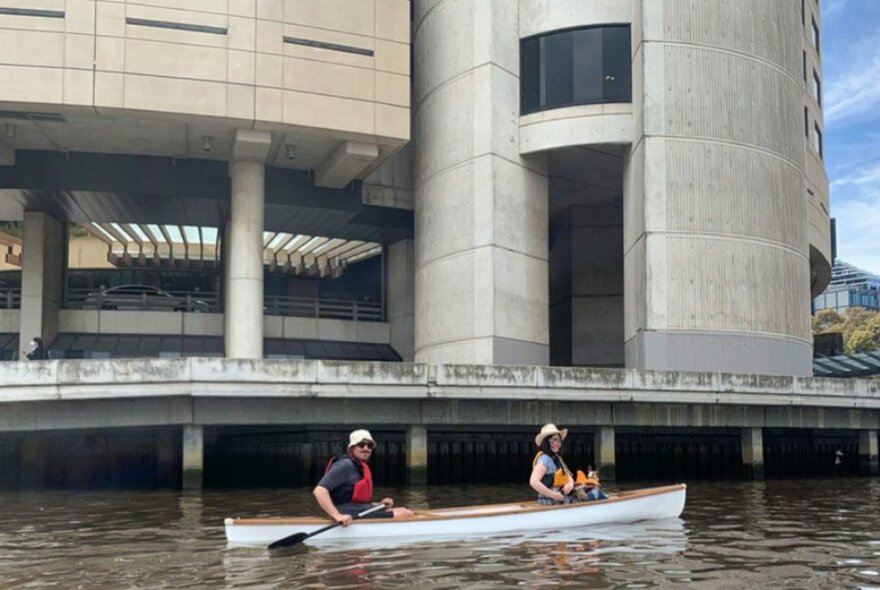 Two people in a canoe on the Yarra River with a large concrete building visible behind them. 