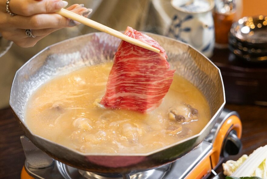 A hand holding chopsticks holding a slice of wagyu beef as it's lowered in to a hot pot. 