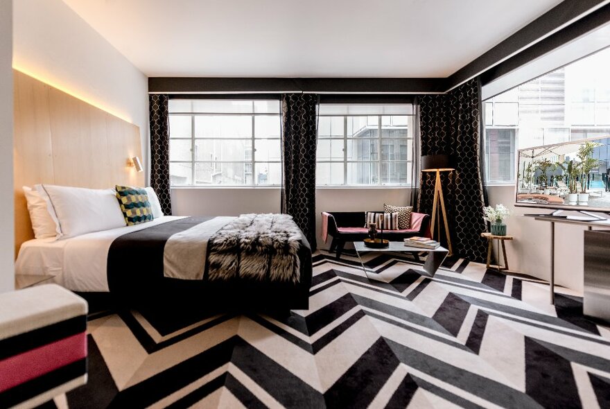 A room at the Adelphi Hotel showing geometric patterned carpet, a king-sized bed with silver, white and black bedding, and dark curtains framing large windows.