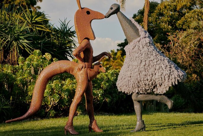 Two people in a garden setting, one in a sculptural kangaroo costume, the other in an emu costume.