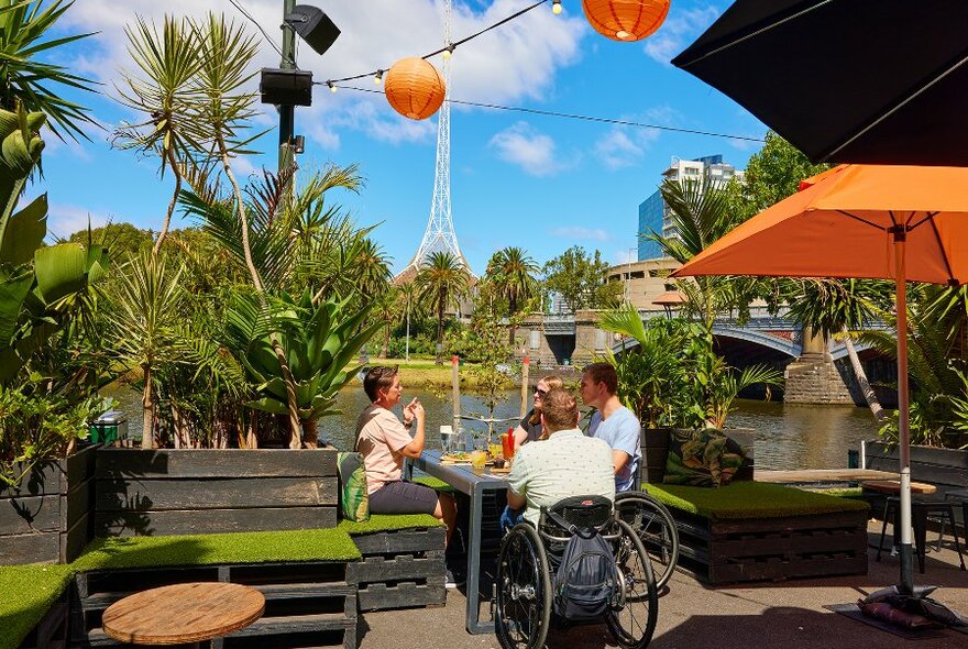 A group dining beside the river at an outdoor bar with orange umbrellas. Two are sitting in wheelchairs. 