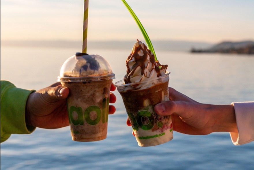 Two hands 'cheersing' plastic cups of yogurt with green spoons sticking out, in front of body of water.