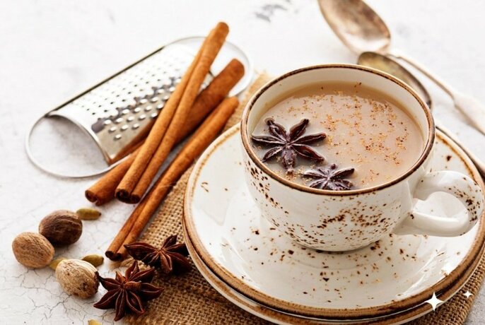 Milky chai (tea) in a cup with cinnamon sticks, star anise and whole nutmegs resting on the table alongside.