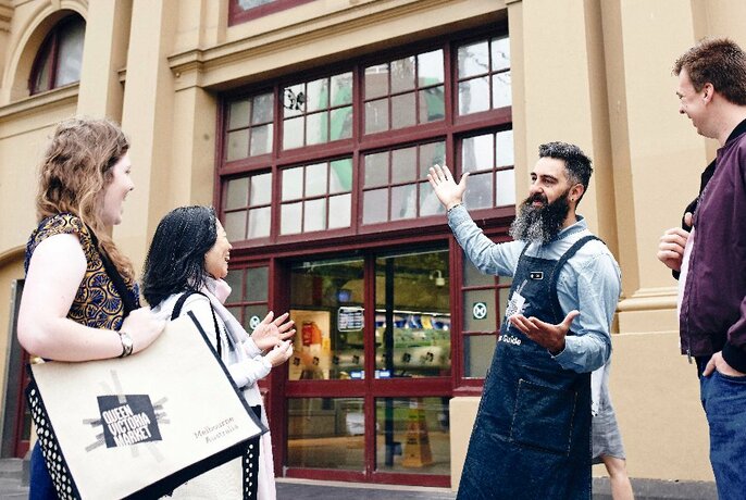 A bearded tour leader with tour participants, pointing at the entrance to Queen Vic Market.