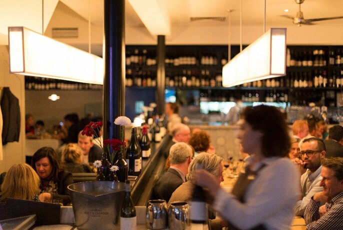 View across busy restaurant, with wine shelving at rear, and waiter in foreground.