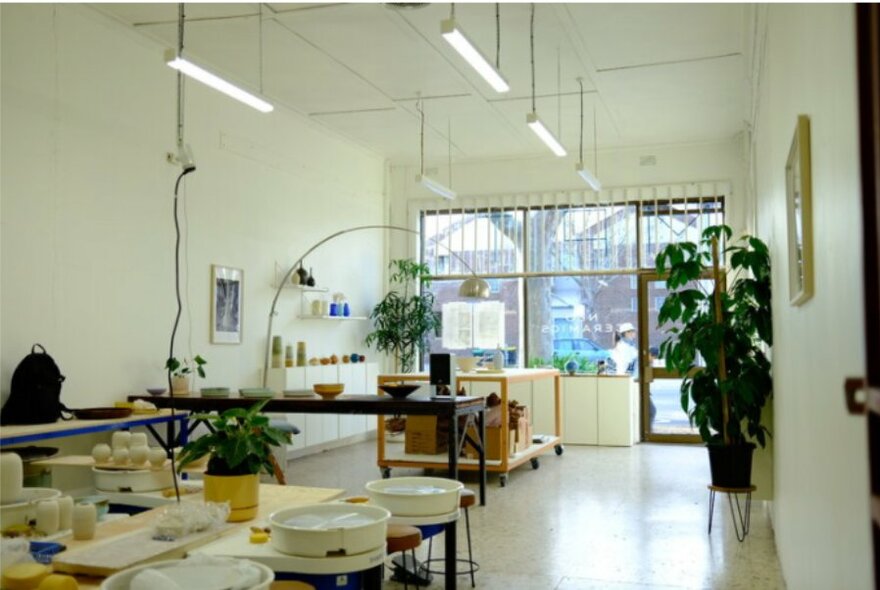 A brightly lit studio shop with white walls and neon lights, a table loaded with white ceramics in the foreground, plants and displays closer to the window and front door.