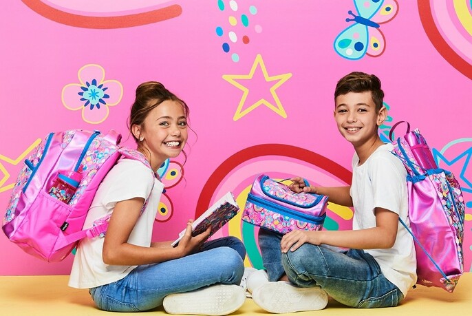 Two children with colourful backpacks, girl holding a notebook, boy holding a lunch box, against pink coloured background.