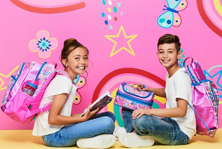 Two children with colourful backpacks, girl holding a notebook, boy holding a lunch box, against pink coloured background.