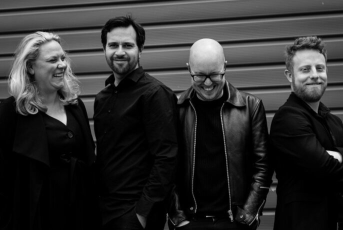 Black and white group portrait of the four members of Syzygy Ensemble; three men and one woman, standing in front of a wall.