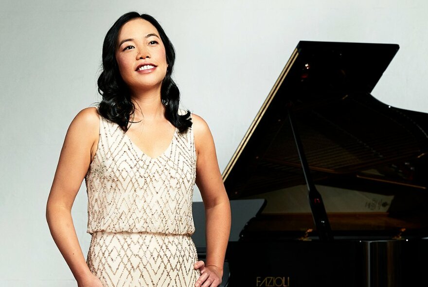 Pianist Andrea Lam in evening wear in front of a black grand piano and a white background.