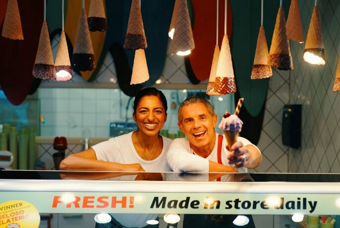 Smiling staff leaning over the glass service counter of a gelato shop holding out a gelato in a cone.