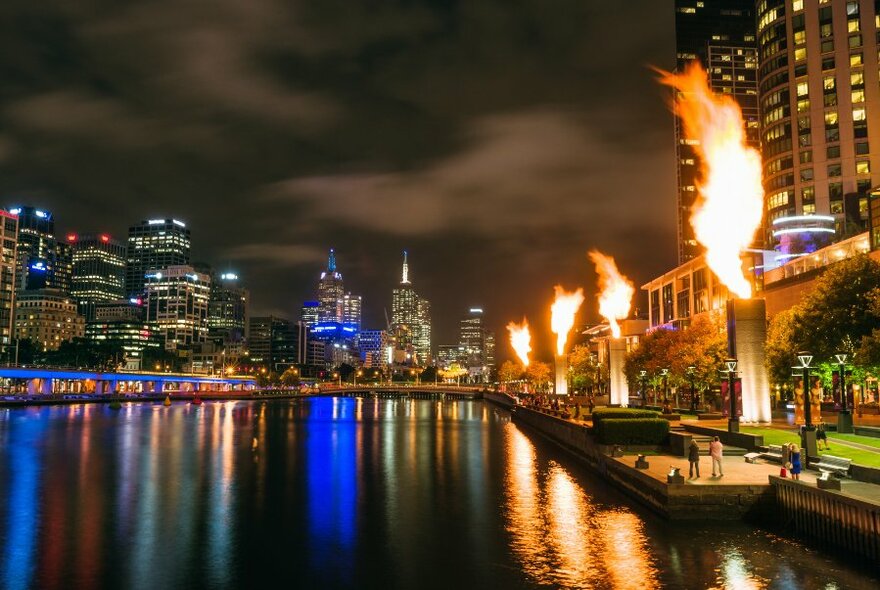 The lights of Melbourne city skyline taken from the Yarra River at night, with flames leaping into the air from a display.