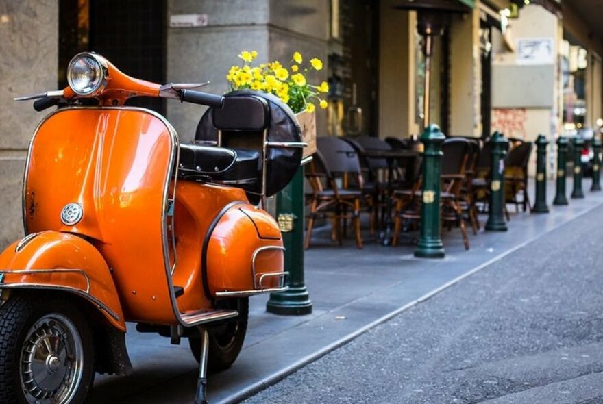 An orange Vespa parked on a sidewalk in a city laneway with cafe tables and chairs in the background.