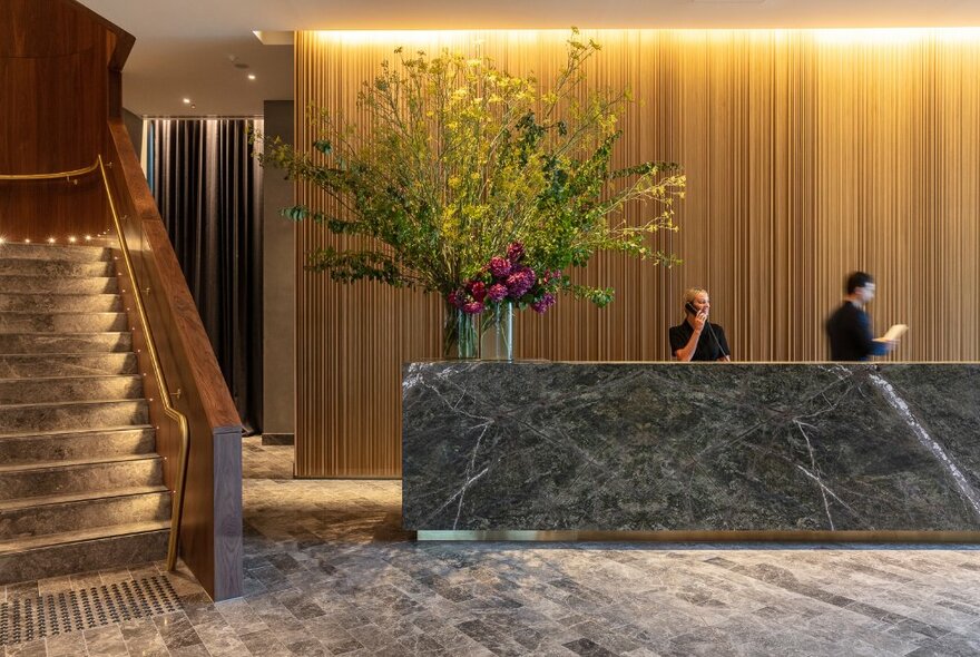 Elegant and spacious foyer of Next Melbourne hotel, showing a large marble check-in counter with an oversize floral arrangement on the desk, and a staircase to the left.
