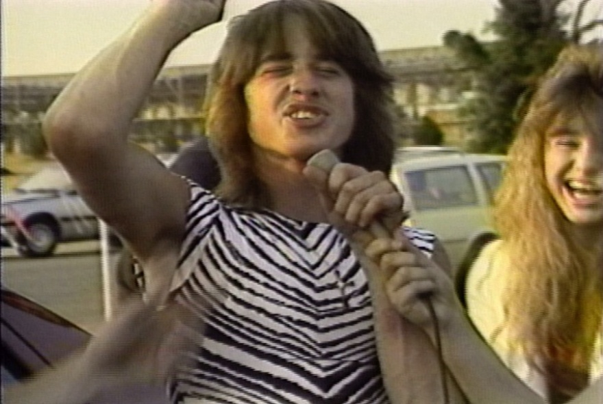 A still from a 1980s video of a young man with long hair, raising his fist in the air and talking into a microphone; outdoors.