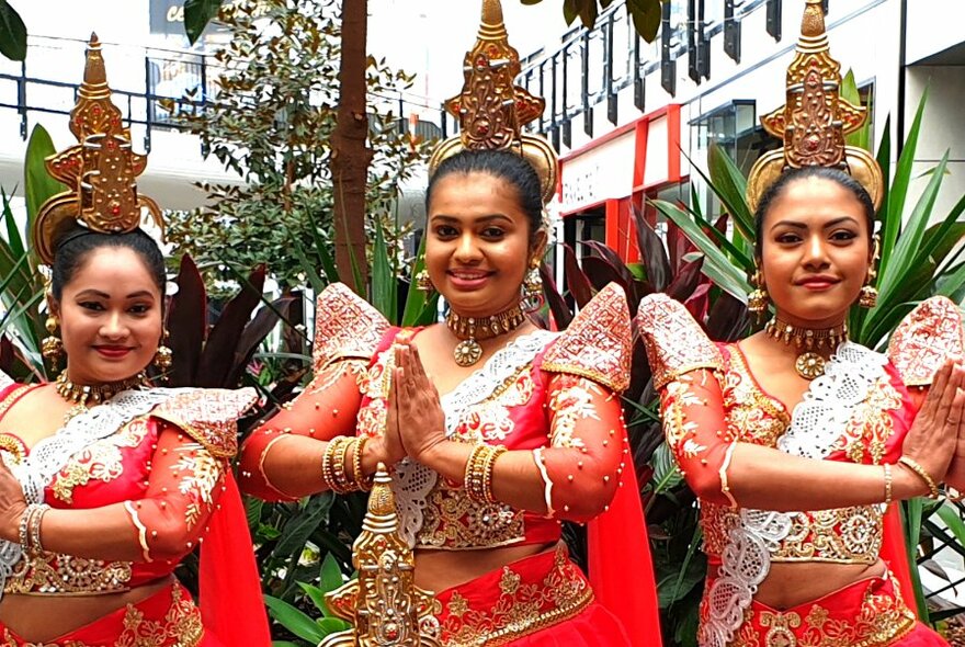 Three women in red and gold Sri Lankan dancing costumes, smiling at camera and standing in a  pose with their hands pressed together.