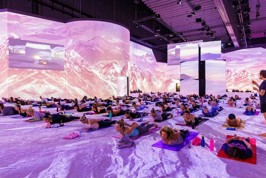 Purple, pink and white lit up room with numerous people in rows doing yoga on mat.